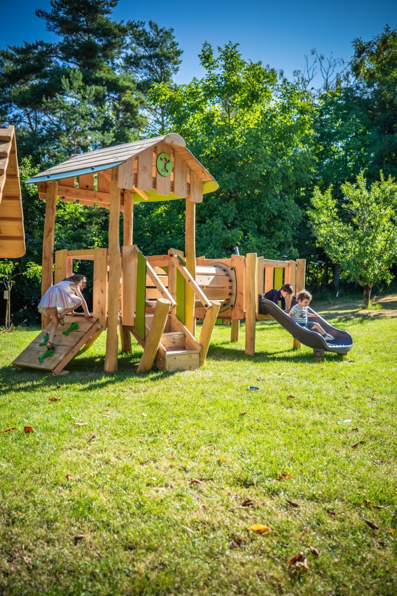 Peuters spelen op een houten speeltoestel met klimhelling, kruiptunnel en glijbaan in een groene buitenruimte.